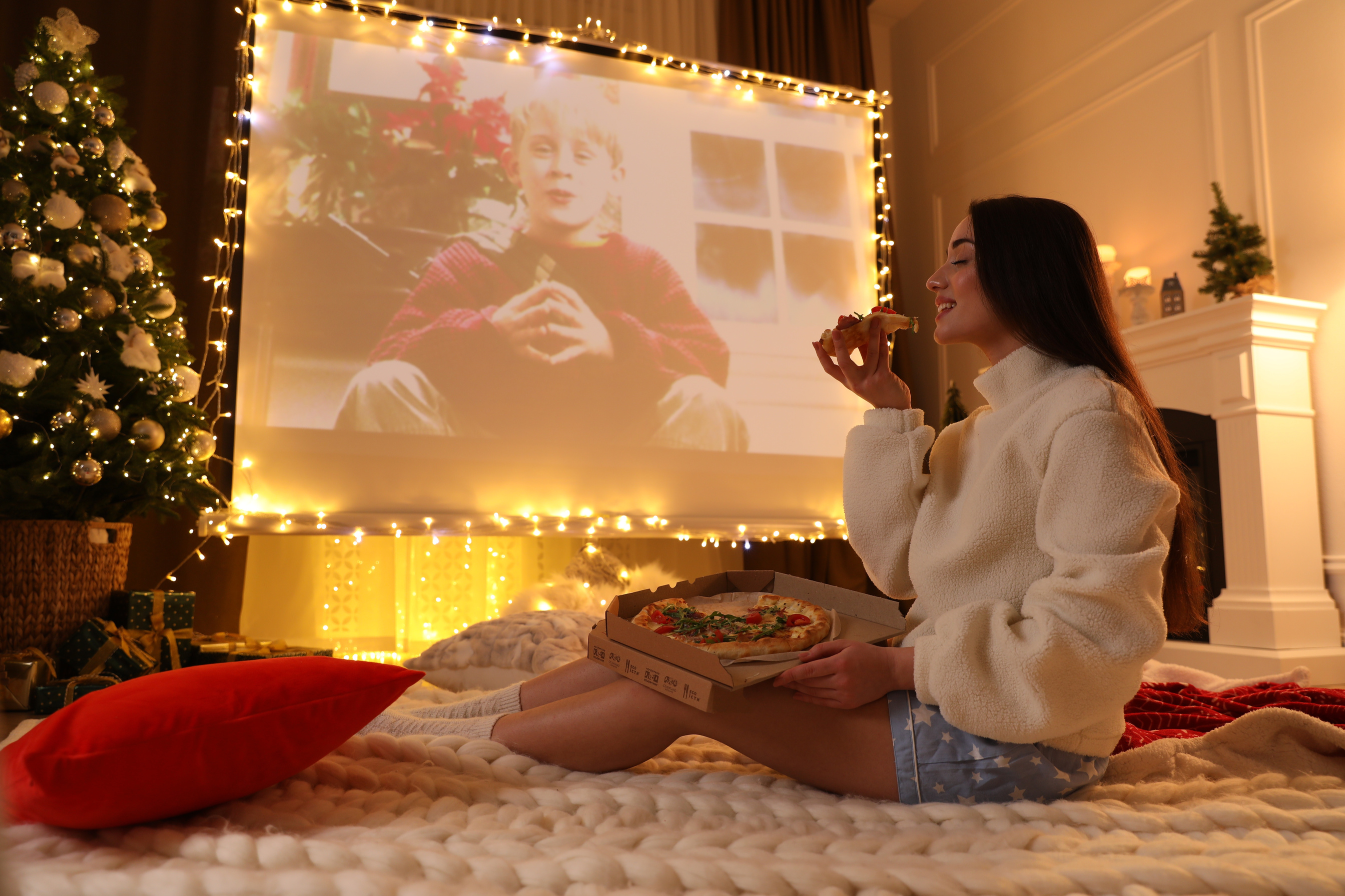 MYKOLAIV, UKRAINE - DECEMBER 24, 2020: Woman Watching Home Alone Movie via Video Projector in Room. Cozy Winter Holidays Atmosphere